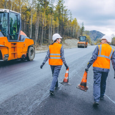 Two workers in orange vests work on a highway in the mountains.