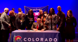 Governor Polis joins Black leaders to sign bill on a stage with a bright blue background.
